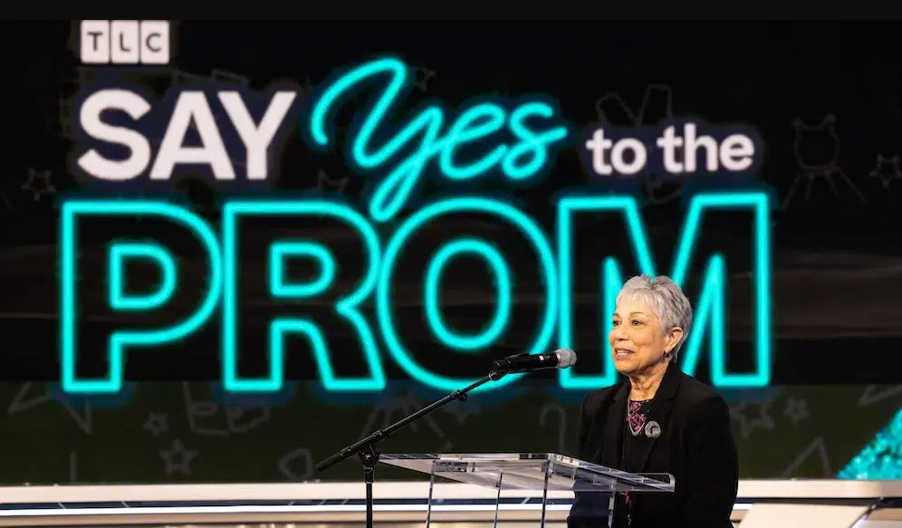A woman speaking in front of the words "Say Yes to the Prom"